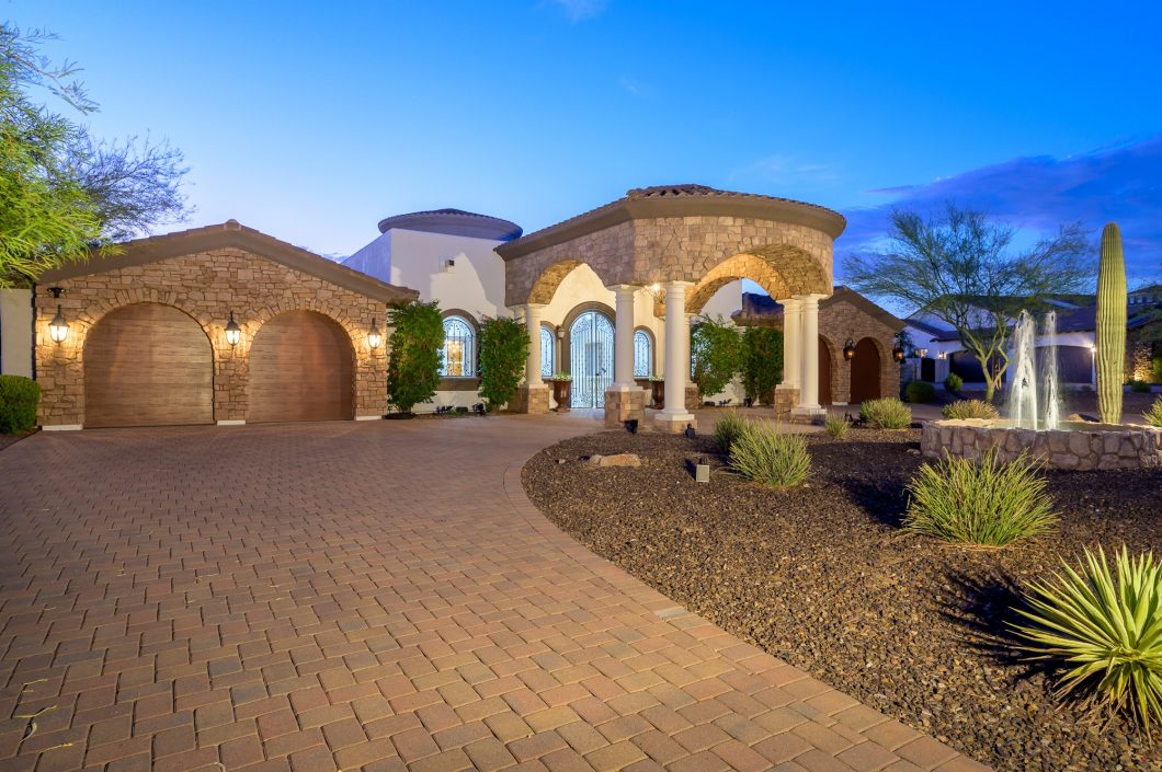 Mediterranean-style luxury home with a stone exterior, arched columns, and a fountain in the desert landscape, featuring a horseshoe driveway and well-lit architectural details.