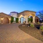 Mediterranean-style luxury home with a stone exterior, arched columns, and a fountain in the desert landscape, featuring a horseshoe driveway and well-lit architectural details.