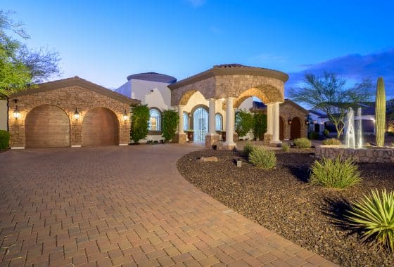 Mediterranean-style luxury home with a stone exterior, arched columns, and a fountain in the desert landscape, featuring a horseshoe driveway and well-lit architectural details.